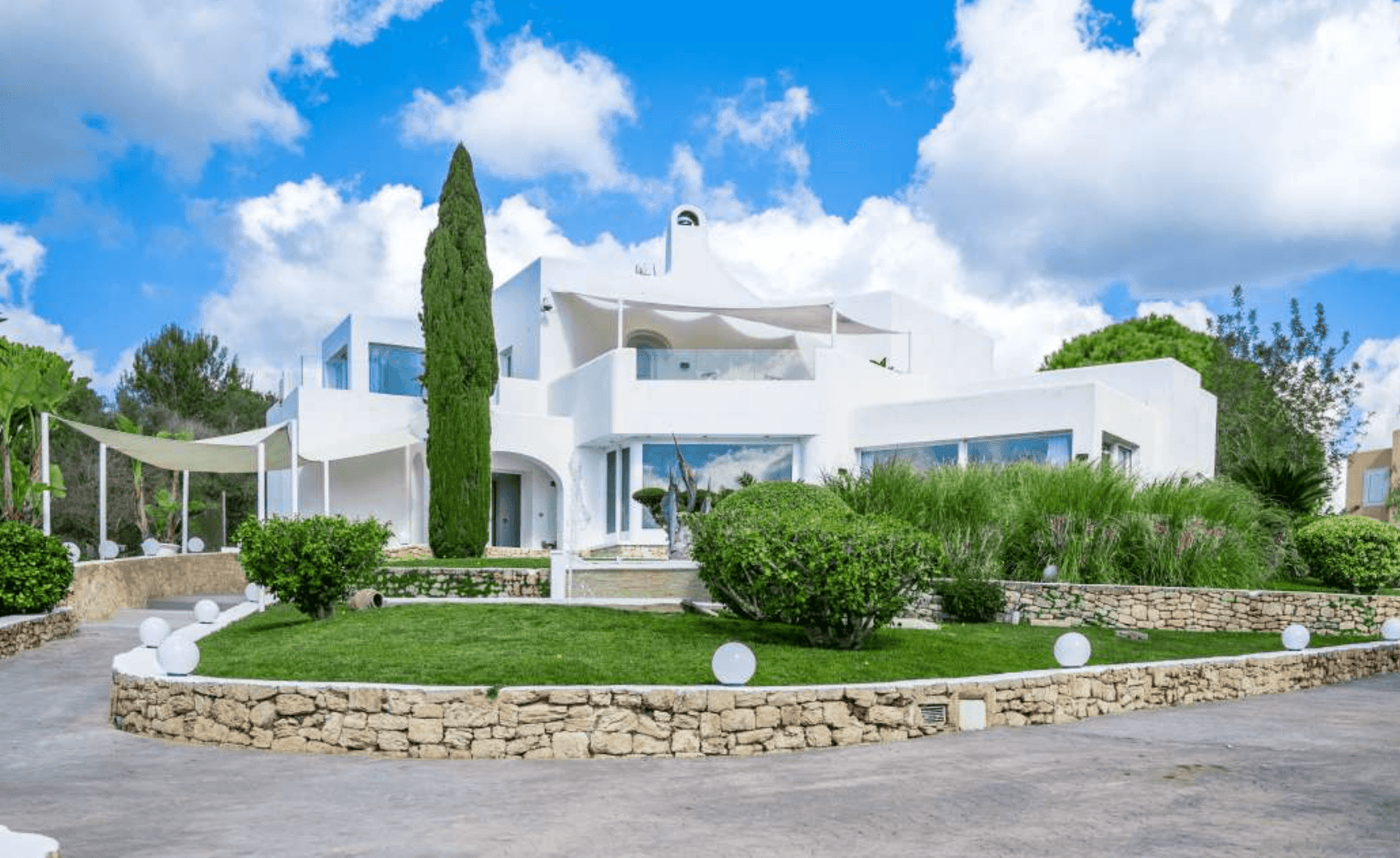 Modern white villa with large windows, surrounded by greenery and stone landscaping under a blue sky.